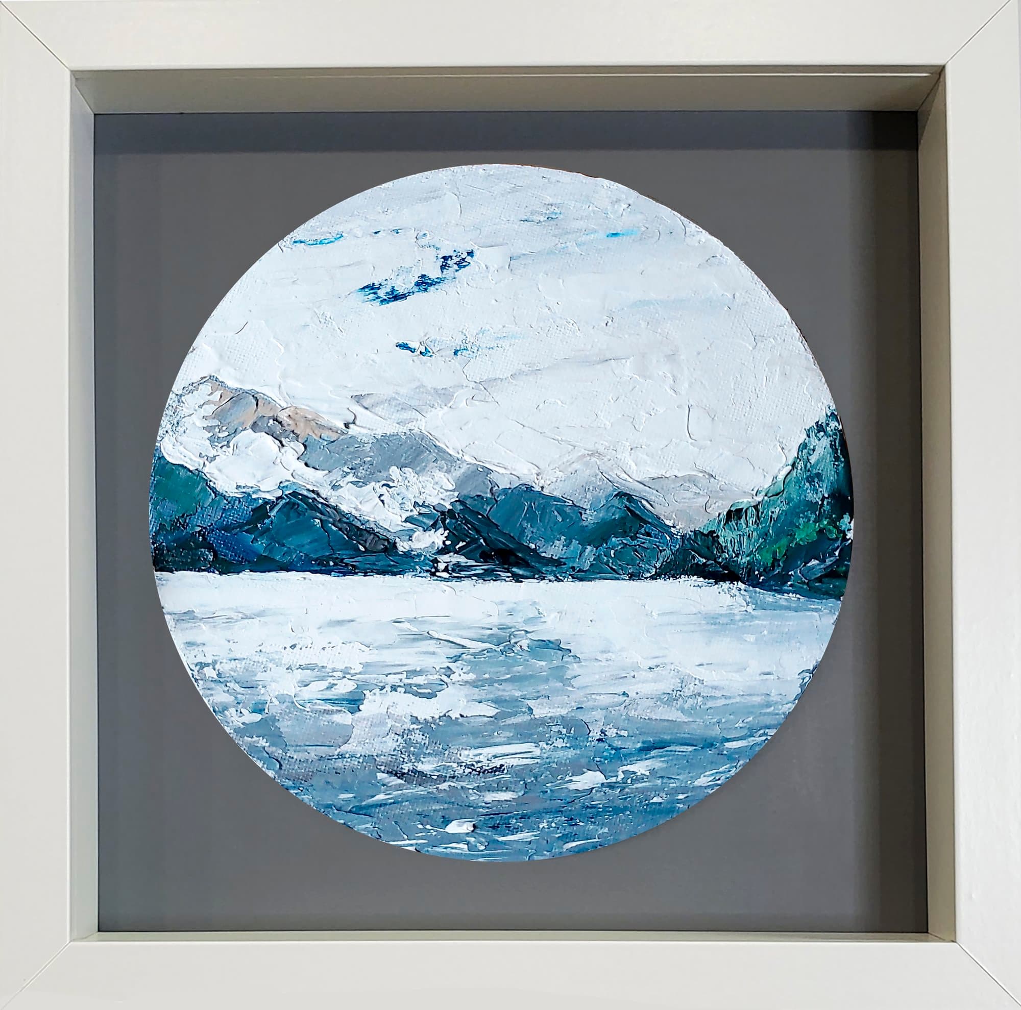 Round painting in a white frame showing mountains and blue lake water